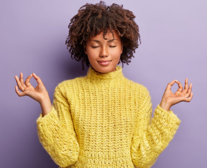 People, peace and calmness concept. Black young female stands in mudra posture, practices yoga, meditates indoor, enjoys peaceful atmosphere, releases stress, reaches nirvana, closes her eyes